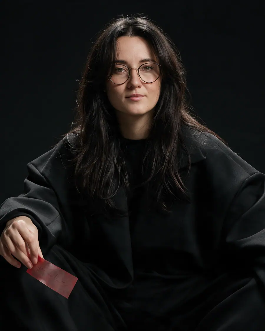 Catherine Tskho seated in a dark studio, holding a small red note card.